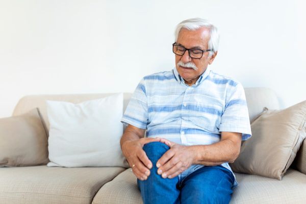 Elderly man sitting on a sofa at home and touching his painful knee